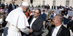 John seated in his power wheelchair shaking hands with Pope Leo.